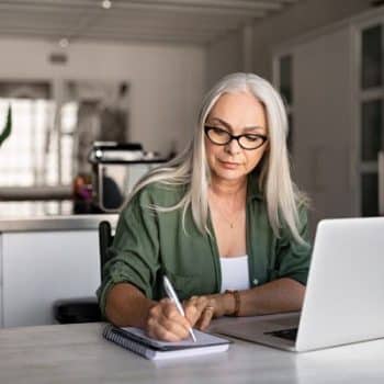 Woman with gray hair and glasses planning a low-carb breakfast menu in a sunlit kitchen, writing in a notebook beside her laptop