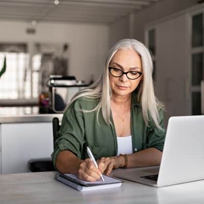 Woman with gray hair and glasses planning a low-carb breakfast menu in a sunlit kitchen, writing in a notebook beside her laptop