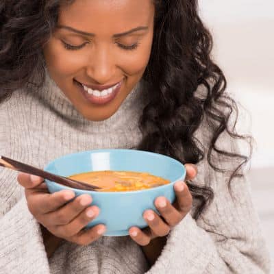Woman in a cream sweater holding a blue bowl of creamy low-carb soup with a wooden spoon