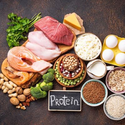 Overhead view of high-protein foods including beef, chicken, salmon, eggs, cheese, beans, and nuts arranged around a small chalkboard sign that reads Protein