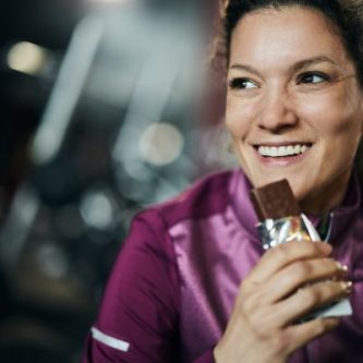 Smiling woman in a purple workout jacket eating a chocolate protein bar at the gym