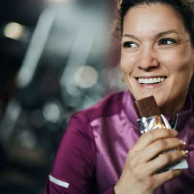 Smiling woman in a purple workout jacket eating a chocolate protein bar at the gym